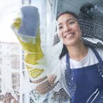 Cheerful young woman smiling while cleaning the window, glass surface using sponge. Housework and housekeeping, cleaning service concept. View through the glass