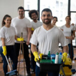 Cheerful young man part of a cleaning team holding a tray with cleaning products all smiling at camera in an office