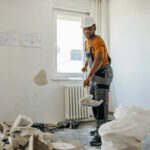 An African American male construction worker in a protective uniform standing in a room, putting away residue with a shovel.